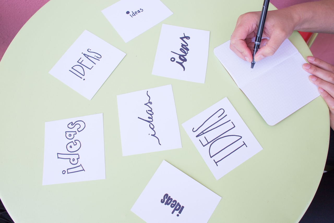 La imagen muestra una fotografía de una mesa con hojas de papel en las que han escrito la palabra “ideas''. Aparece también una mano a punto de escribir en un cuaderno